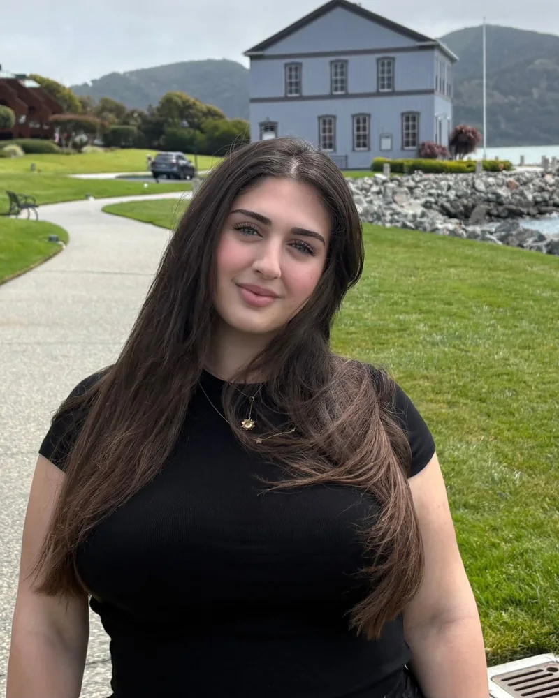 Erva Şengül smiling on a waterfront path, with green lawns, hills and a pale blue house behind her